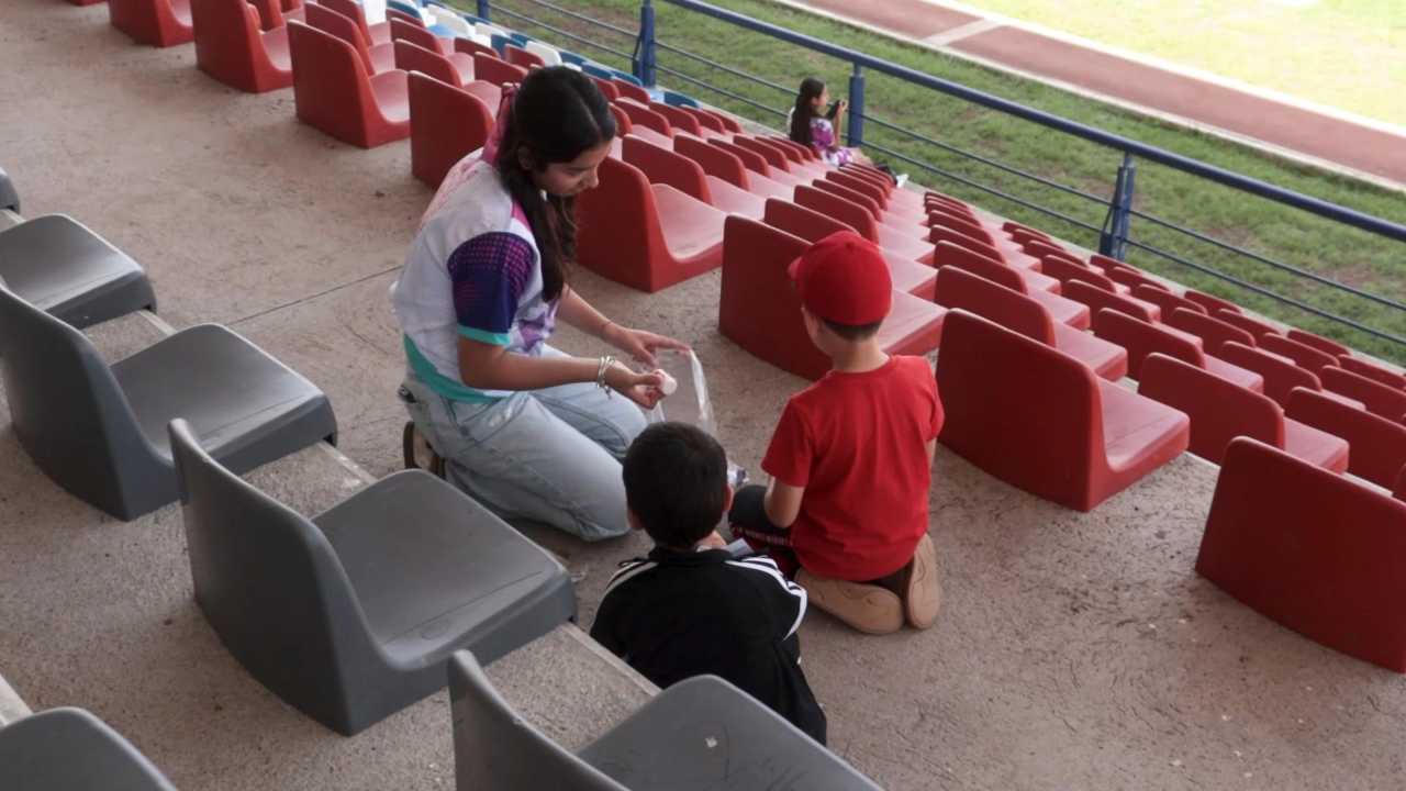 Una de las monitoras asignadas dialoga con dos niños participantes en una zona de sombra de las gradas, como parte de la estrategia de la institución para fomentar una sana convivencia y acercar a las infancias a la vida universitaria. Una de las monitoras asignadas dialoga con dos niños participantes en una zona de sombra de las gradas, como parte de la estrategia de la institución para fomentar una sana convivencia y acercar a las infancias a la vida universitaria.